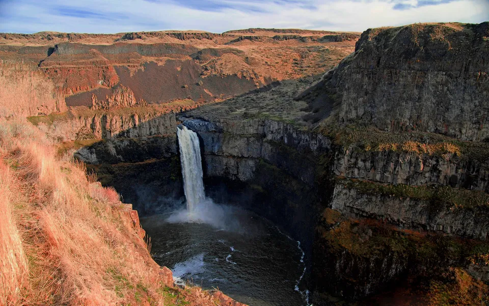 A tall waterfall cascades into a dark pool surrounded by rugged, layered cliffs and dry vegetation.
