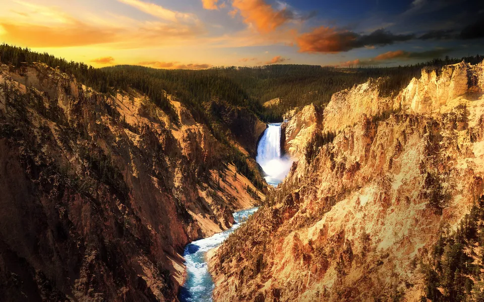 A wide waterfall cascades down a rocky canyon under a dramatic sunset sky.