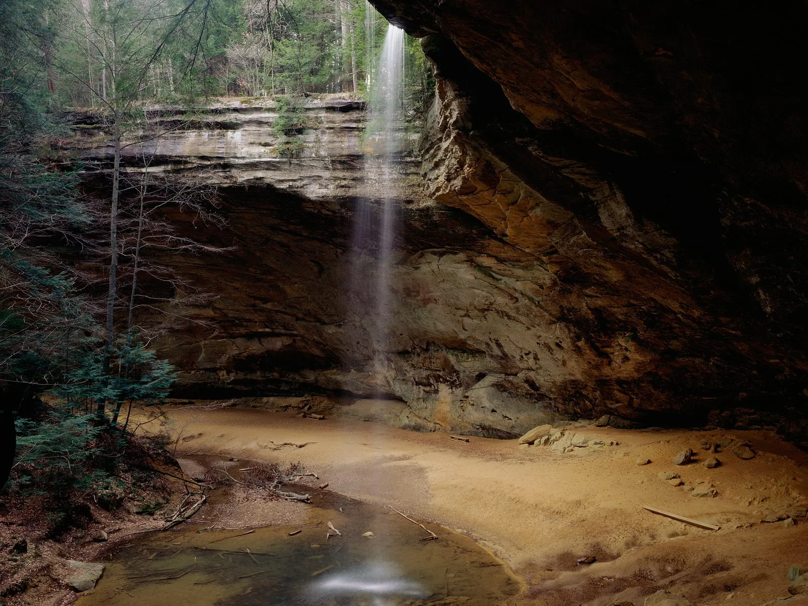 A thin waterfall cascades down a rocky cliff face into a sandy cave area with a small pool of water.