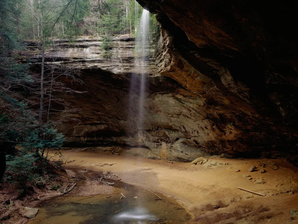 A thin waterfall cascades down a rocky cliff face into a sandy cave area with a small pool of water.