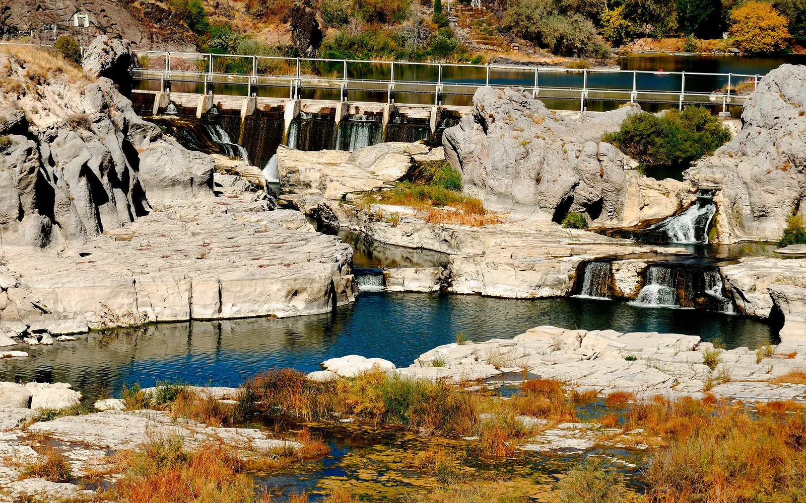 A wide river flows over rocky ledges, with a dam structure visible in the background.