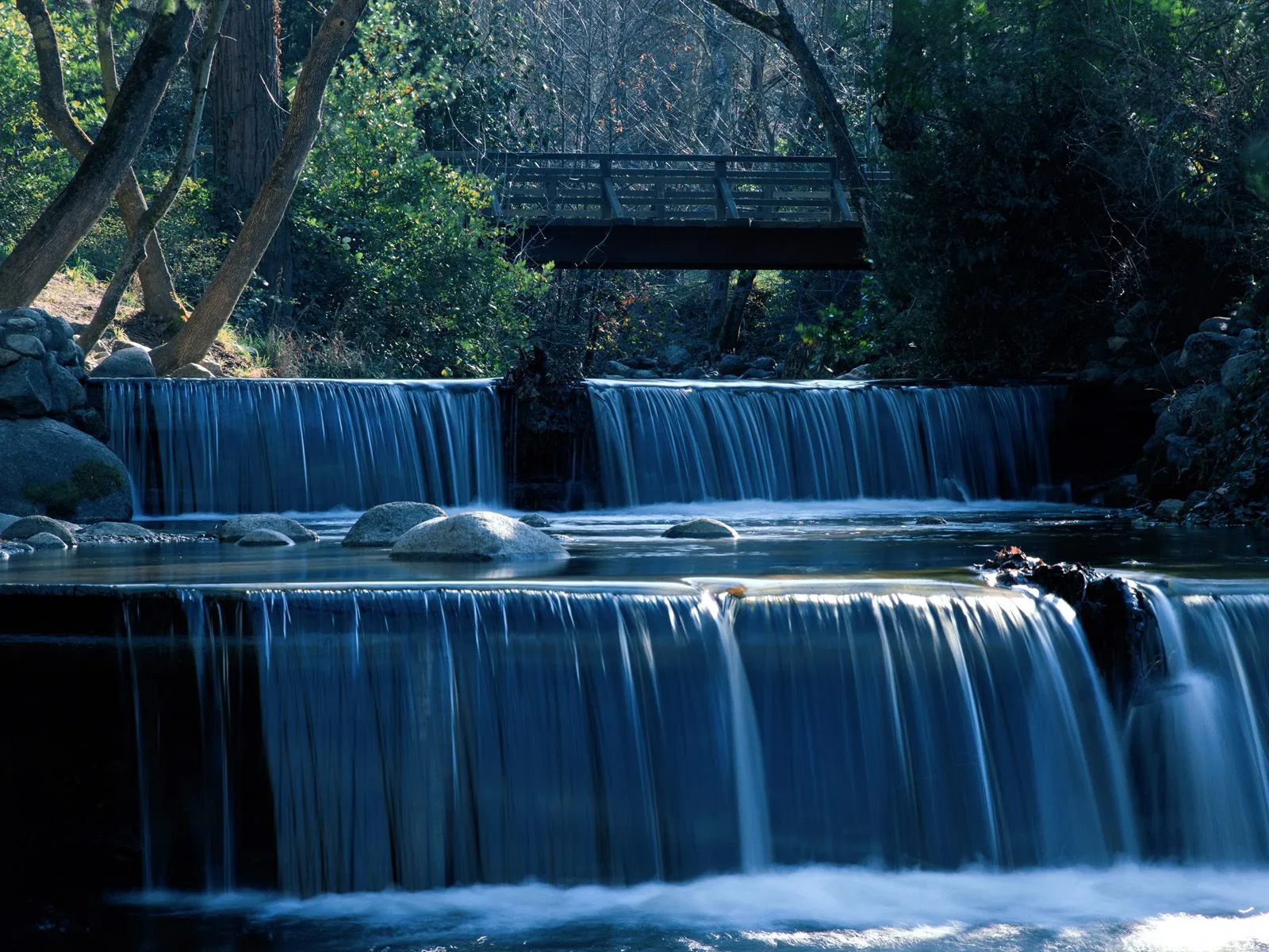 A tiered waterfall cascades over rocks in a forest, with a wooden bridge visible above.