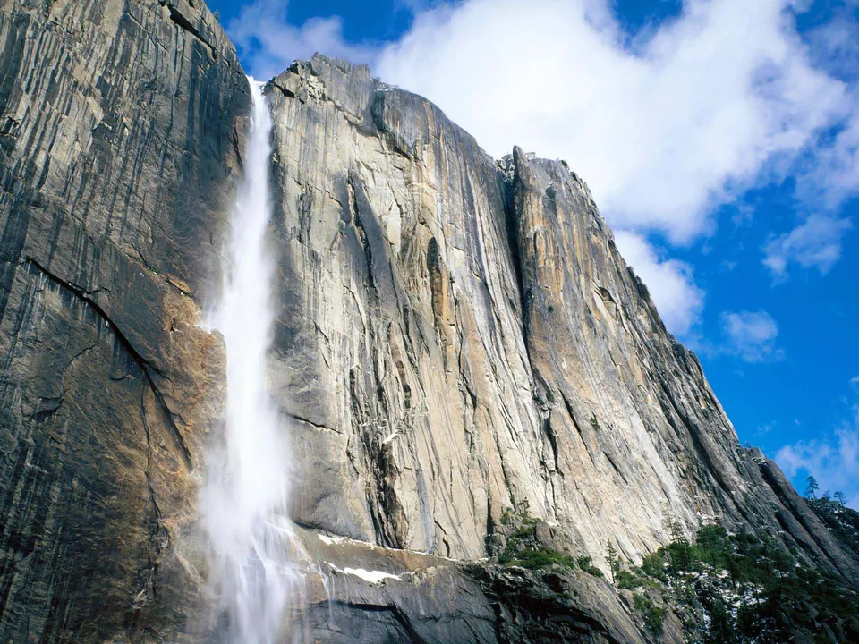 A tall waterfall plunges down a massive, textured granite cliff face under a blue sky with white clouds.