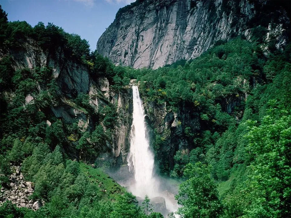 A tall, powerful waterfall plunges down a rocky cliff face surrounded by dense green forest.