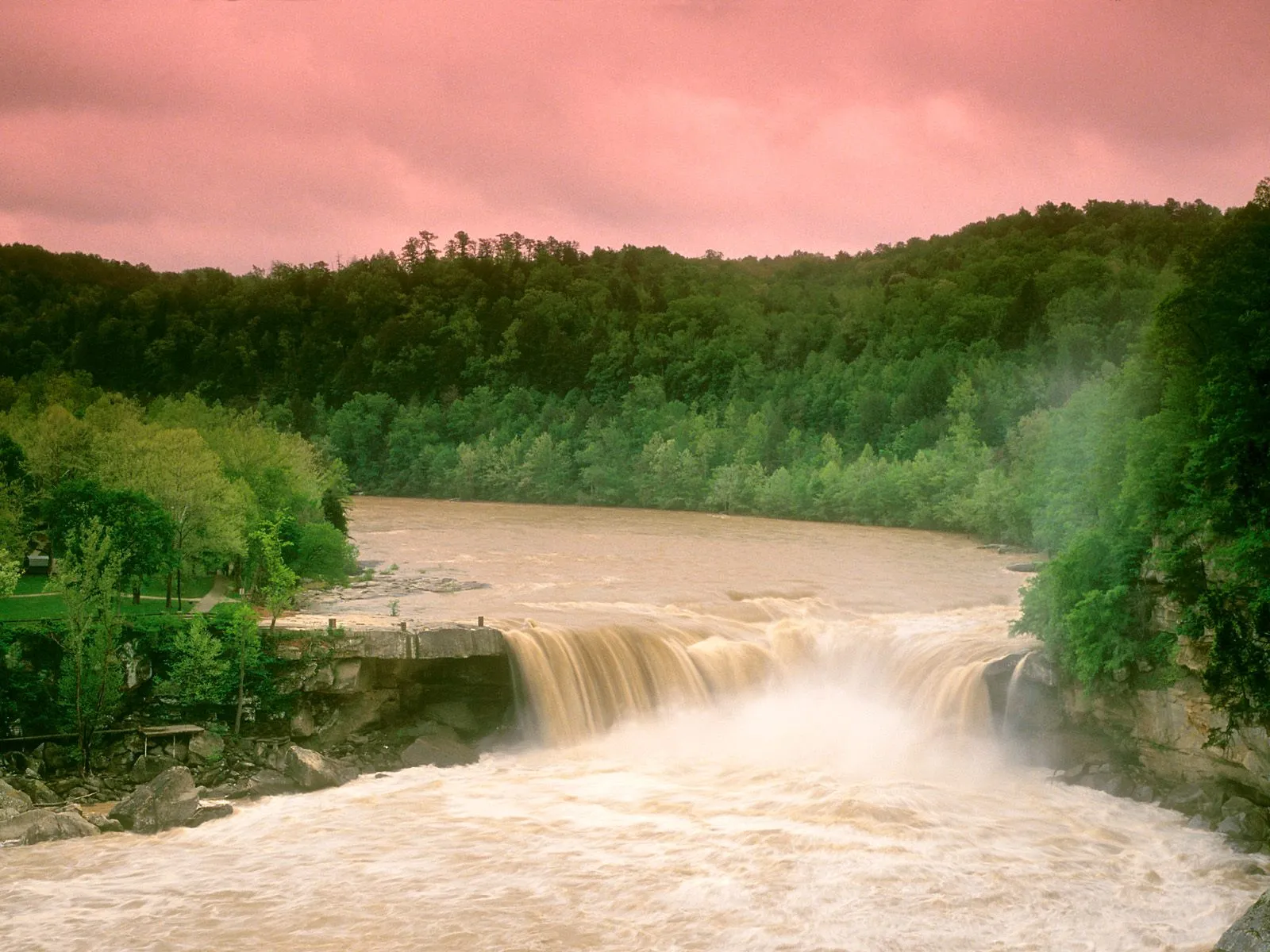 A wide waterfall plunges over a rocky ledge into a turbulent river, surrounded by dense green trees under a pinkish sky.