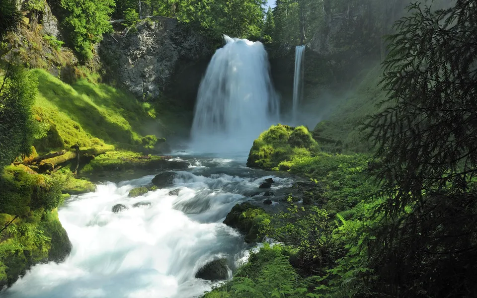 A wide, powerful waterfall cascades down a rocky cliff into a rushing river, surrounded by vibrant green moss and trees.