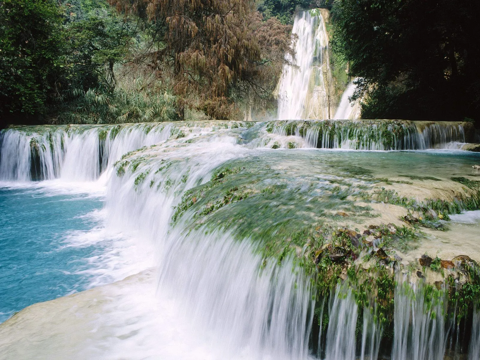 A multi-tiered waterfall cascades over moss-covered rocks into bright blue water, surrounded by lush green foliage.