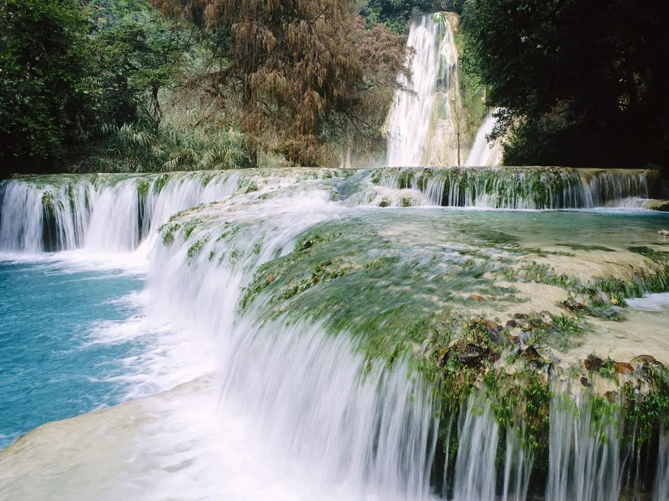 A multi-tiered waterfall cascades over moss-covered rocks into bright blue water, surrounded by lush green foliage.