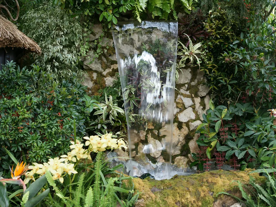 A man-made waterfall cascades down a stone wall surrounded by dense green foliage and yellow orchids.