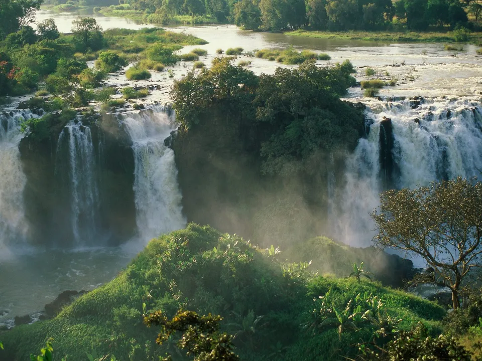 A wide waterfall cascades down rocky cliffs surrounded by vibrant green vegetation and trees.