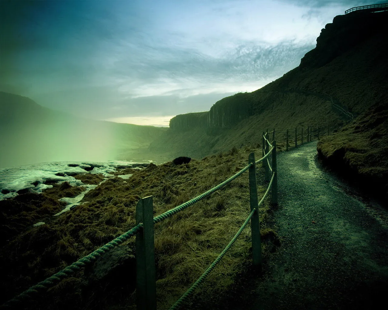 A gravel path with a rope railing winds along a grassy cliff overlooking a misty, icy landscape.
