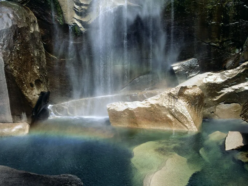 A rainbow forms in the mist of a waterfall cascading into a clear pool surrounded by rocks.