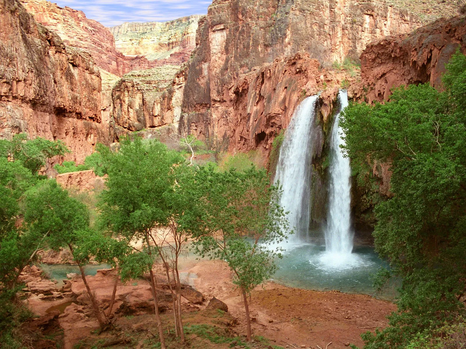 A wide waterfall cascades into a turquoise pool surrounded by red rock cliffs and green trees.