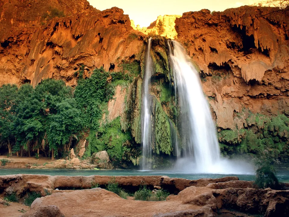 A wide waterfall plunges down a sheer, orange-red cliff face covered in green moss and vegetation into a turquoise pool below.