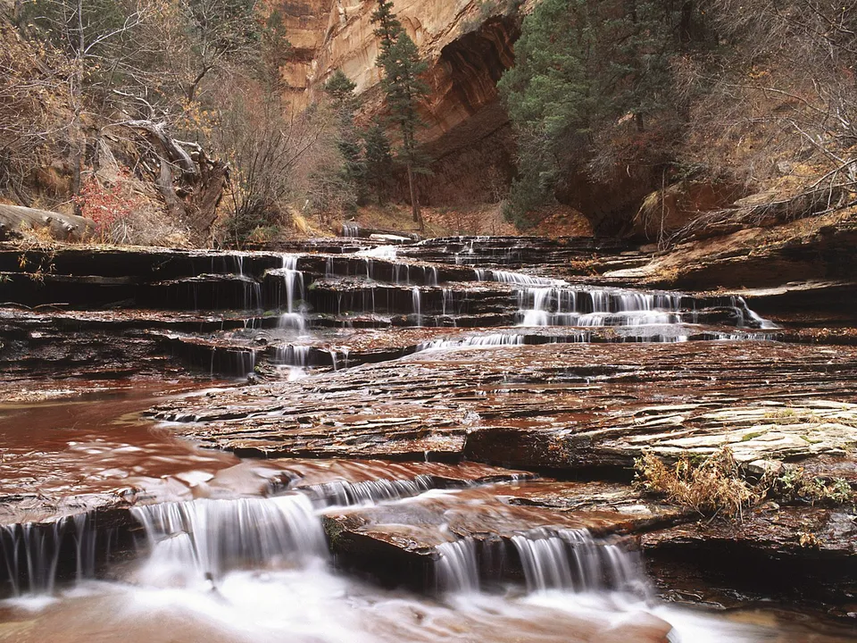 A multi-tiered waterfall flows over flat, layered red rocks in a canyon.