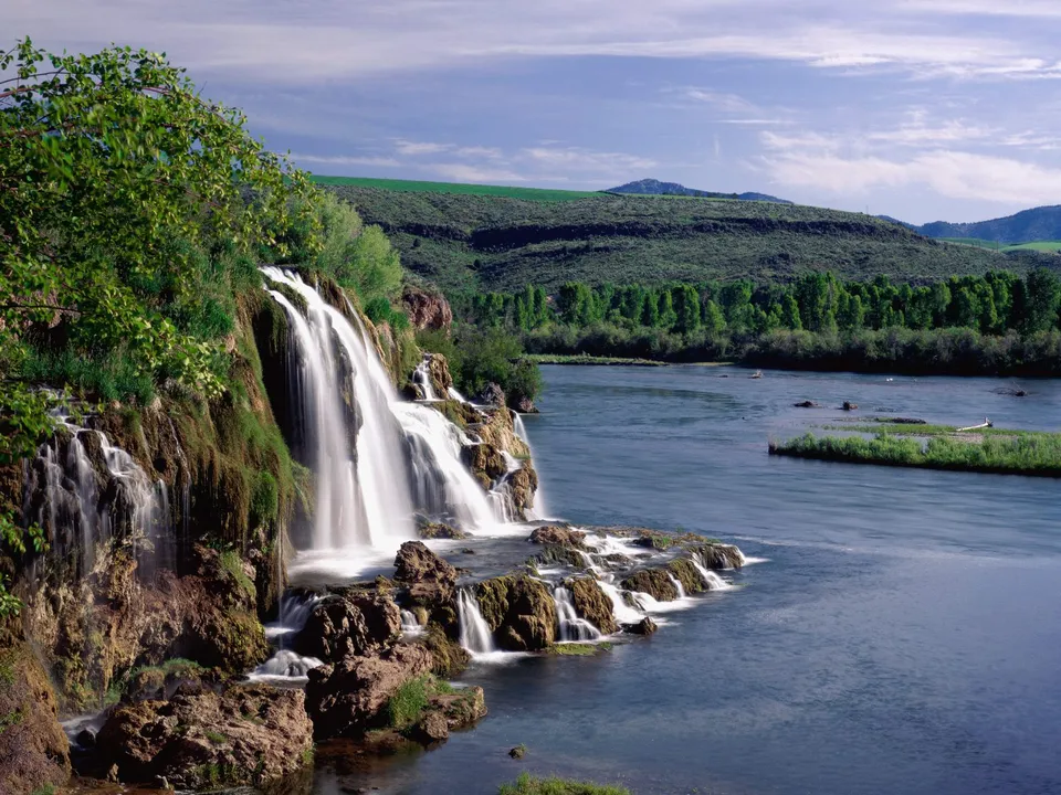 A wide, multi-tiered waterfall flows over mossy rocks into a calm river, with green hills and trees in the background.