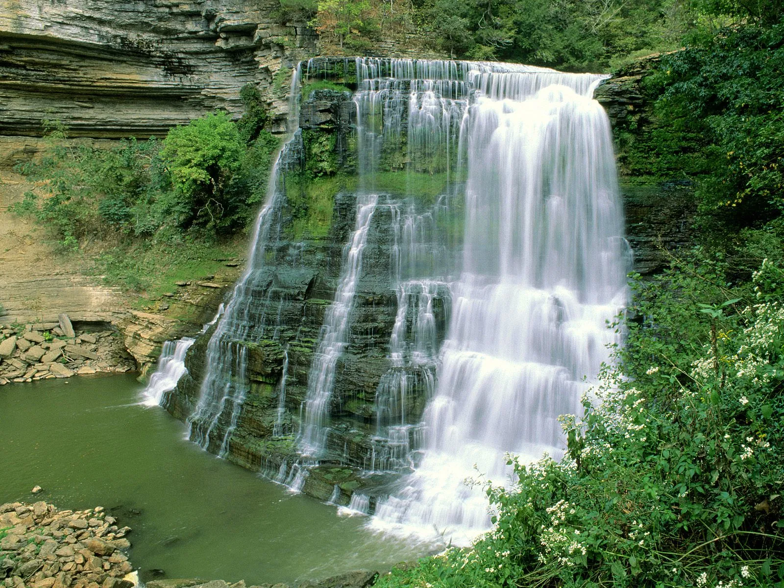 A wide, multi-tiered waterfall cascades down a rocky cliff face into a green pool below, surrounded by lush vegetation.
