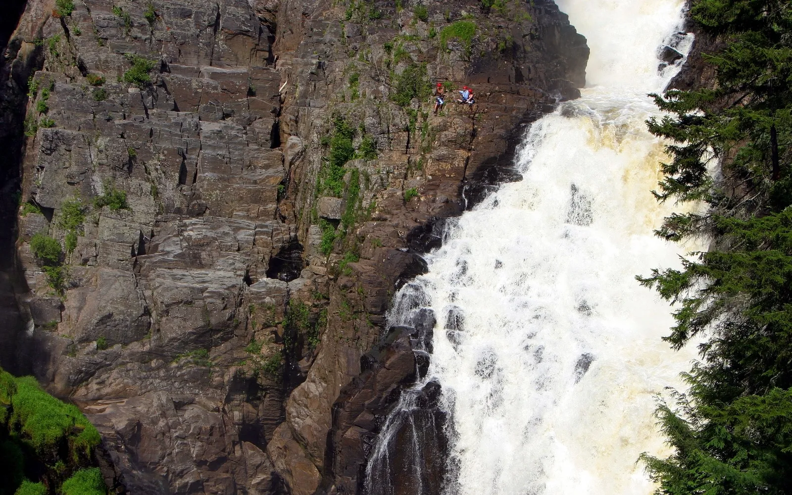 Two people rope jumping from a cliff edge next to a large, powerful waterfall.