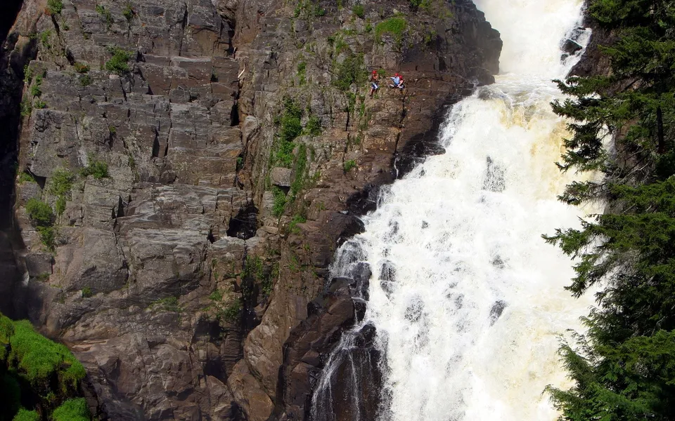 Two people rope jumping from a cliff edge next to a large, powerful waterfall.