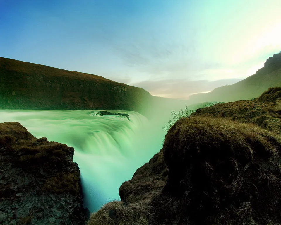 A wide waterfall with milky green water flows over a cliff, surrounded by rocky, grassy terrain and distant hills under a pale sky.
