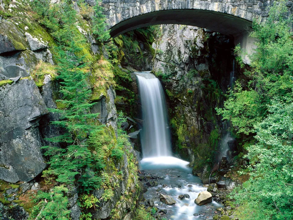 A waterfall flows down a rocky cliff under a stone arch bridge, surrounded by lush green vegetation.