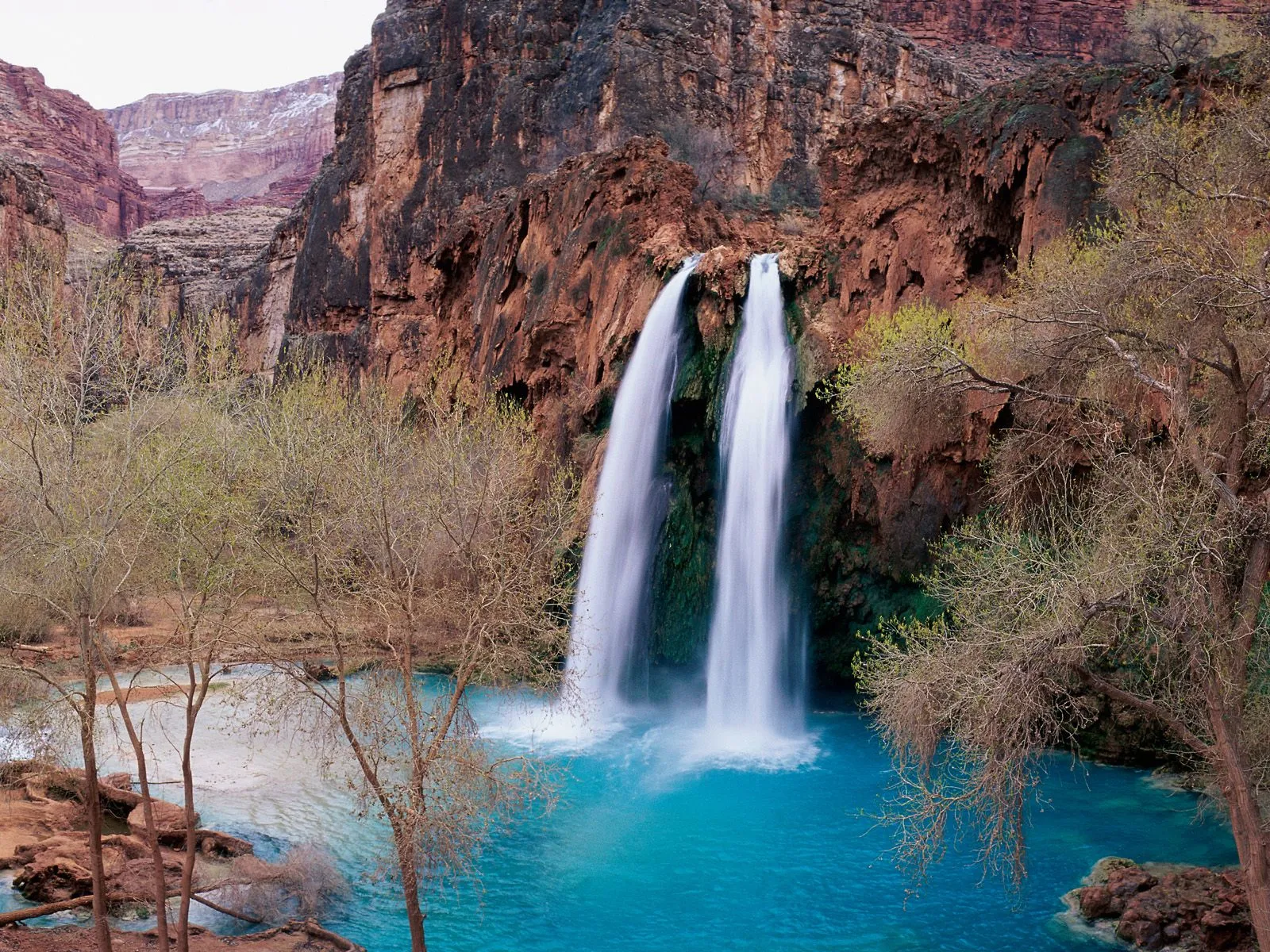 A double waterfall plunges into a vibrant turquoise pool surrounded by red rock cliffs and sparse trees.