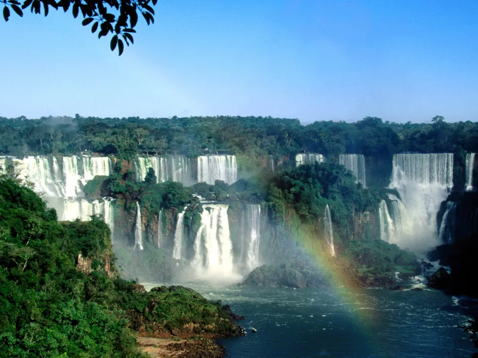 A wide view of multiple waterfalls cascading down lush green cliffs, with a rainbow visible in the mist.