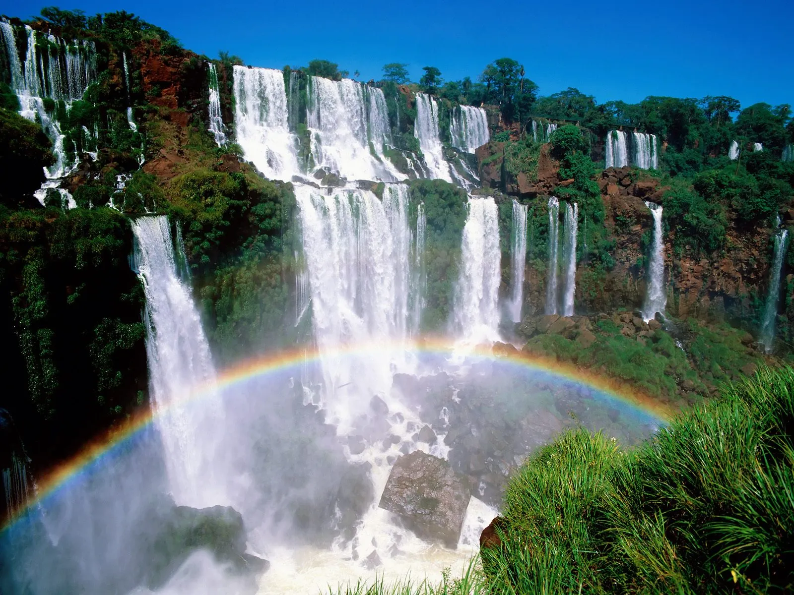 A wide view of numerous waterfalls cascading down a rocky cliff face, with a rainbow visible in the mist.