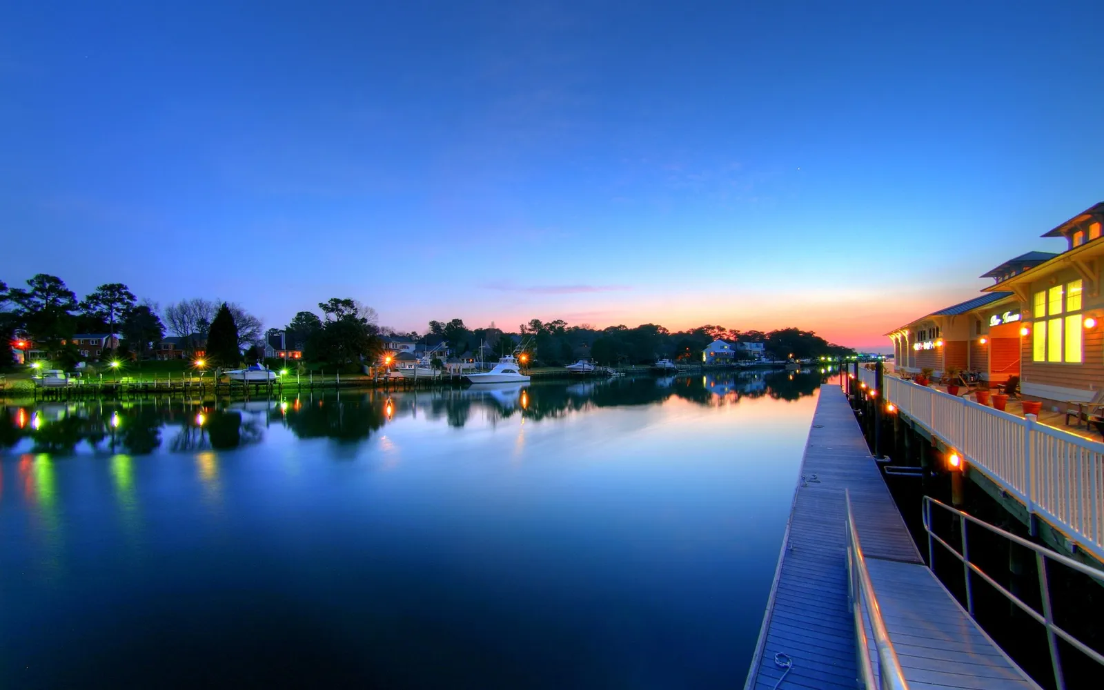 A wooden boardwalk runs alongside a calm river reflecting lights from buildings and trees under a twilight sky.