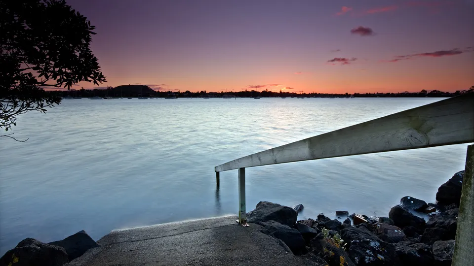 A serene sunset over a calm body of water, with a wooden ramp leading into the water and silhouetted trees on the horizon.