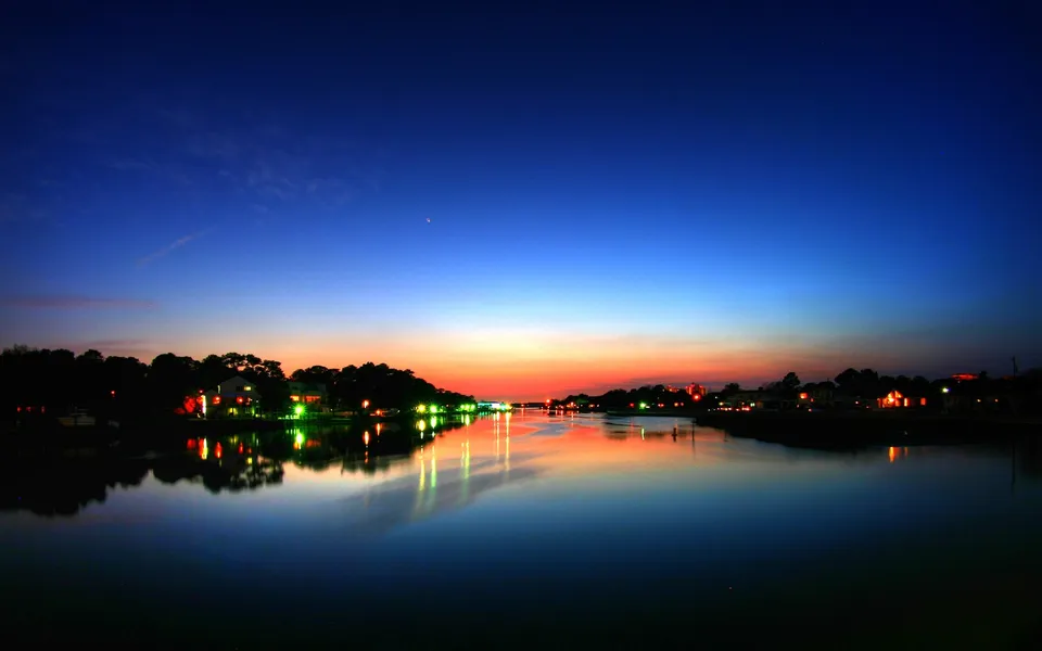 A wide river or bay reflects the colorful twilight sky and illuminated buildings along the tree-lined shore.