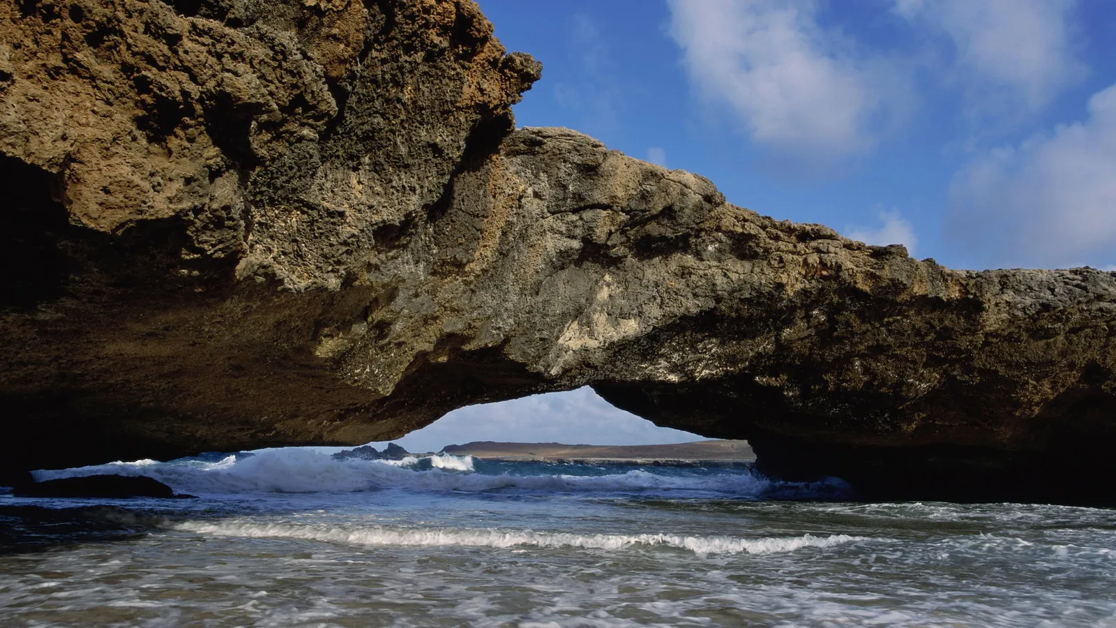 Waves crash through a natural rock arch on a sunny day with blue skies and clouds.