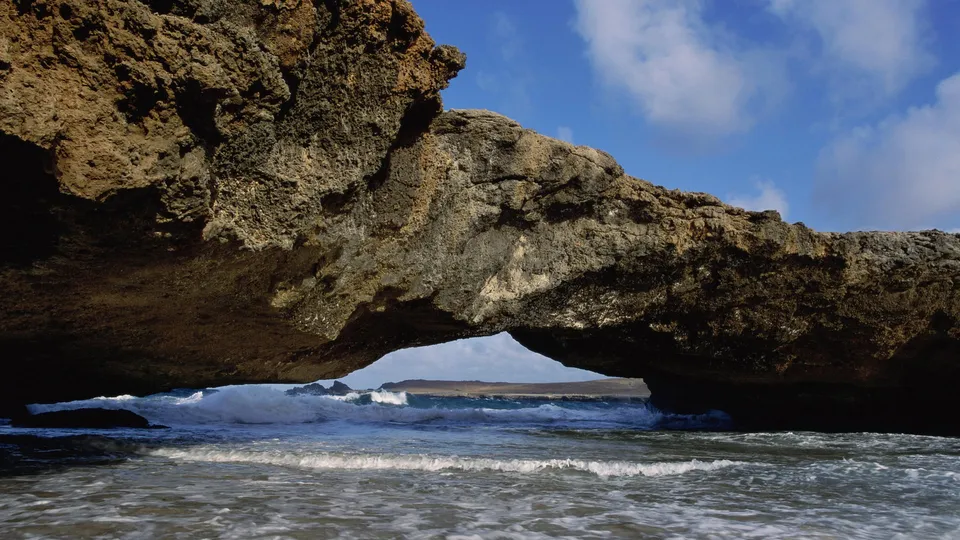 Waves crash through a natural rock arch on a sunny day with blue skies and clouds.