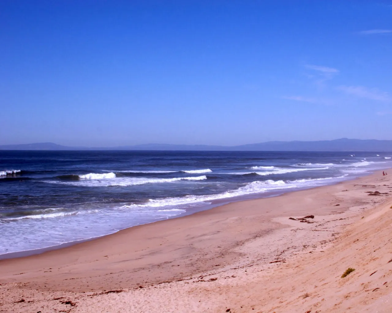 A wide sandy beach with gentle waves rolling onto the shore under a clear blue sky.