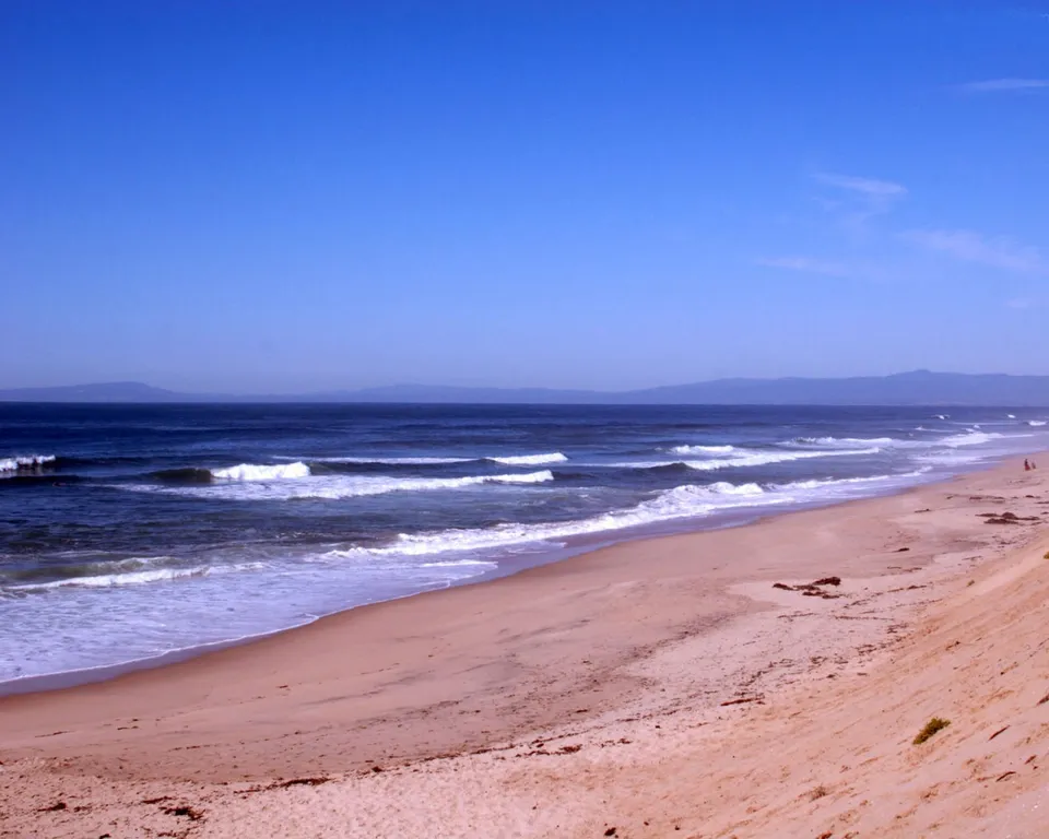 A wide sandy beach with gentle waves rolling onto the shore under a clear blue sky.