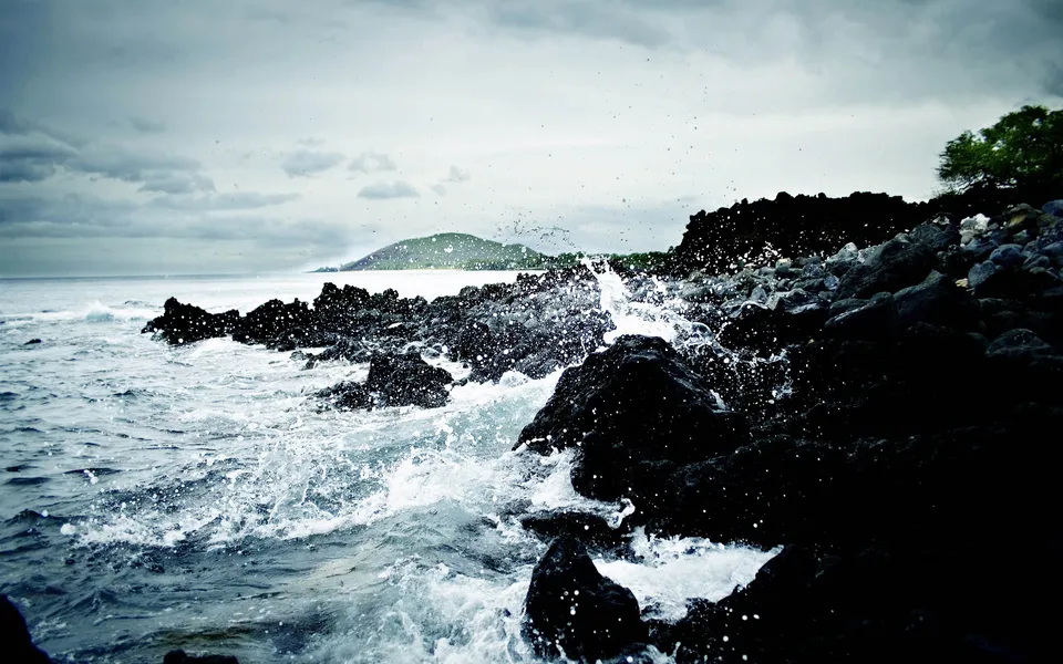 Waves crash against dark, jagged volcanic rocks on a cloudy day, with a distant green island visible.