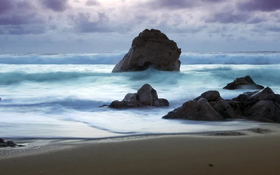 Long exposure shot of waves crashing against dark, jagged rocks on a sandy beach under a cloudy sky.