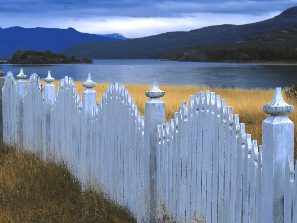 A weathered white picket fence in the foreground with a calm lake and distant mountains in the background.