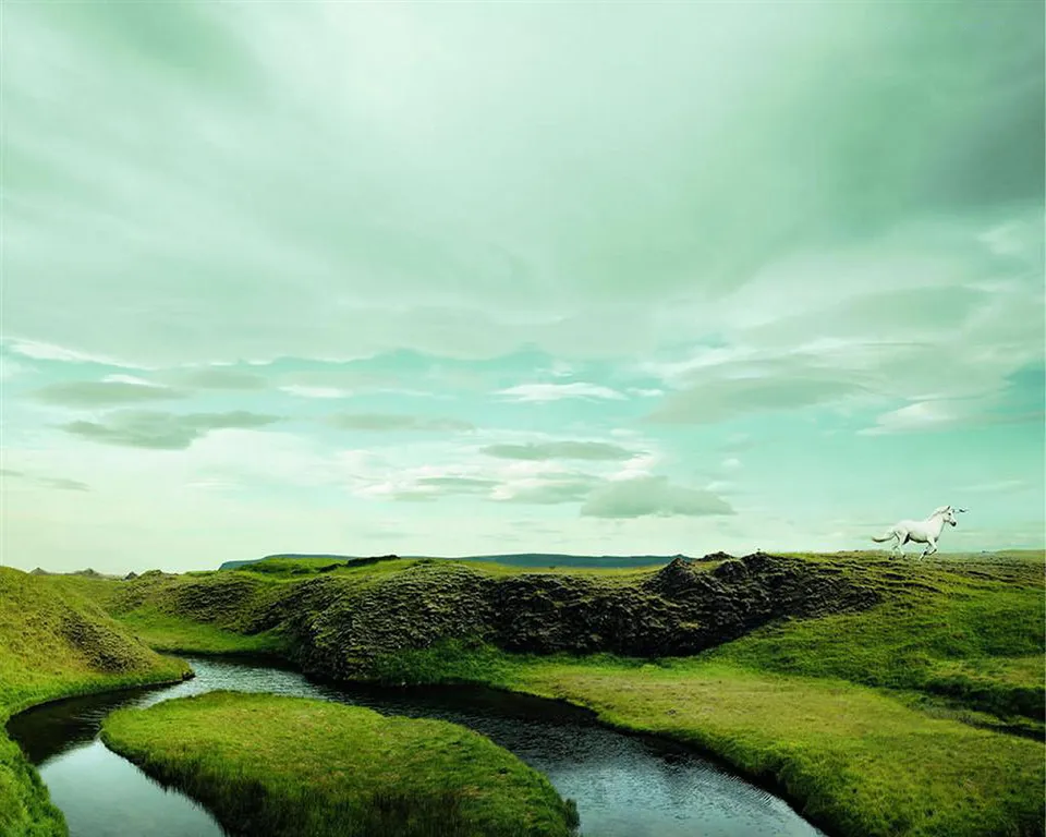 A white horse with a horn runs across a grassy hill next to a winding river under a cloudy sky.