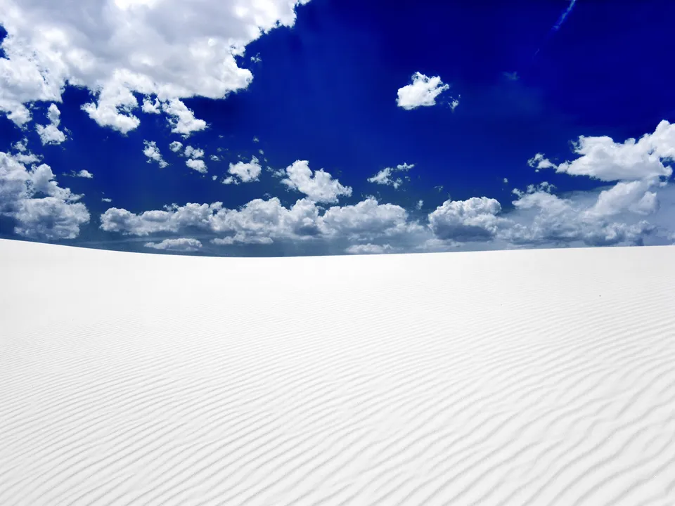 Expansive white sand dunes with rippled textures under a vibrant blue sky with scattered white clouds.