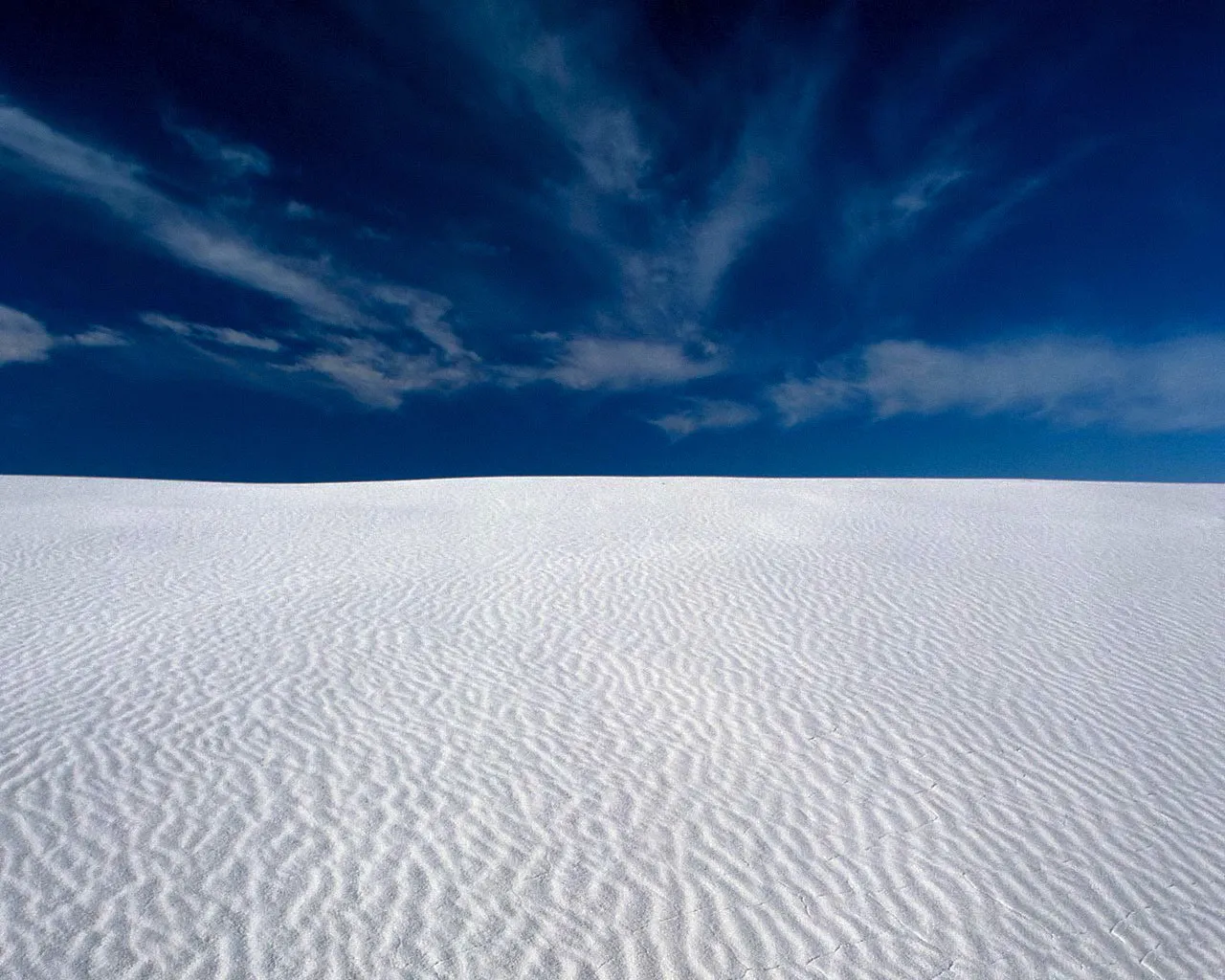 Rippled white sand dunes stretch to the horizon under a vibrant blue sky with wispy clouds.