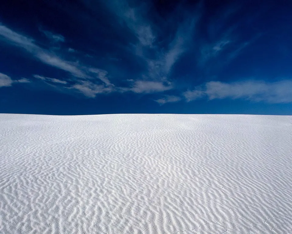 Rippled white sand dunes stretch to the horizon under a vibrant blue sky with wispy clouds.