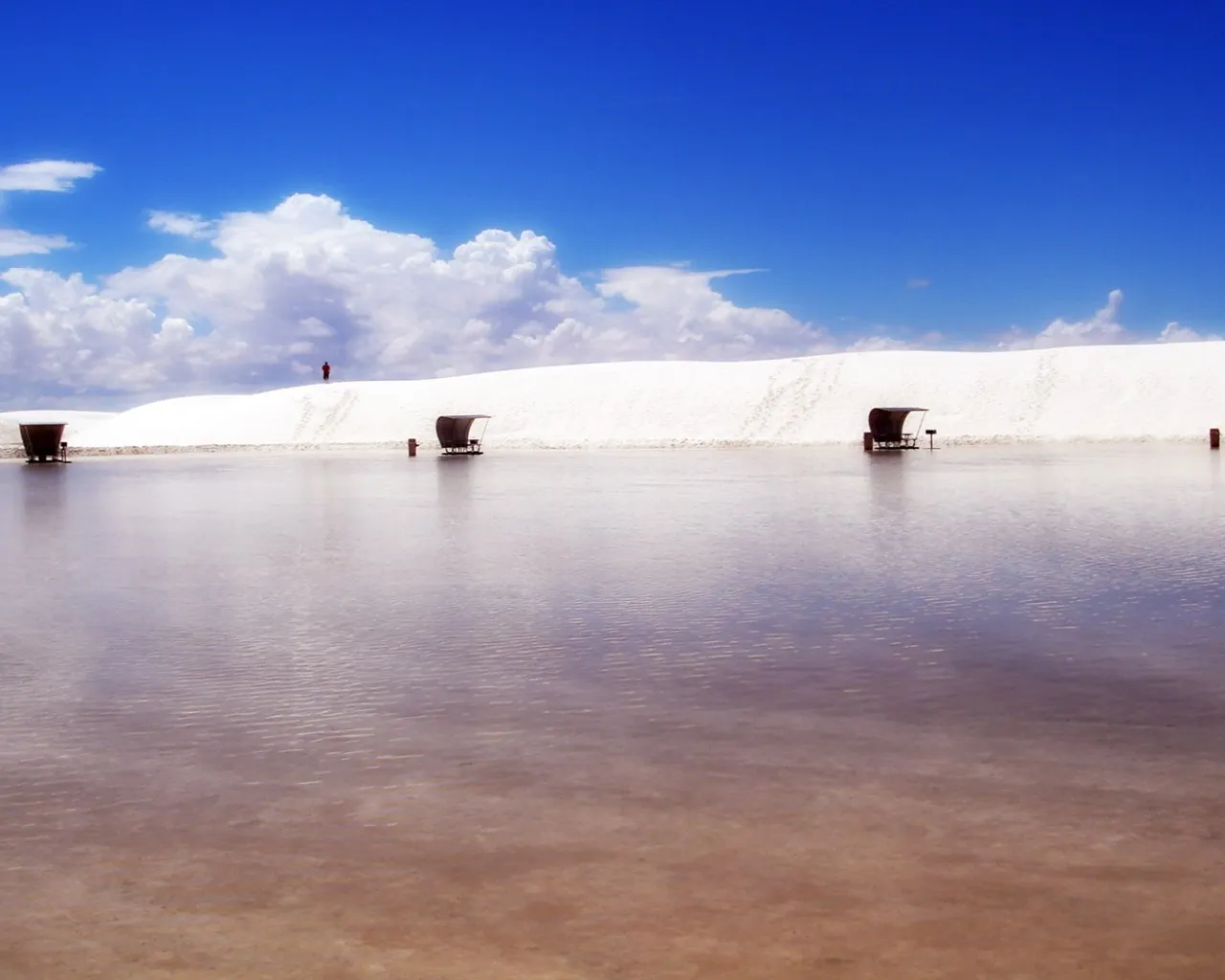 A vast expanse of white sand dunes with a shallow body of water reflecting the blue sky and clouds.