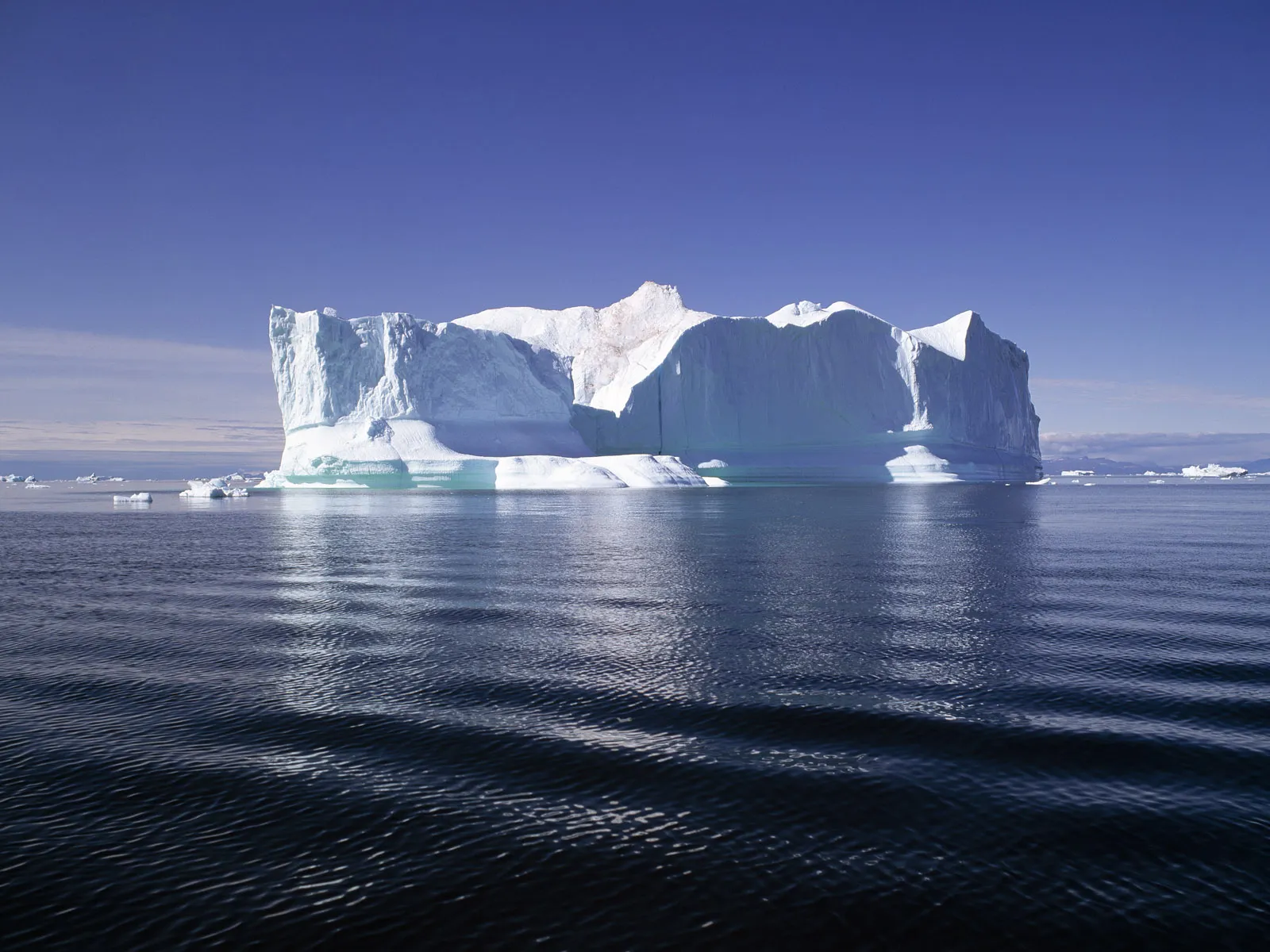 A wide iceberg formation with layered ice sits in a calm, deep blue ocean under a clear sky.