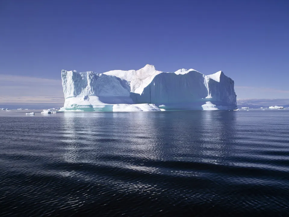 A wide iceberg formation with layered ice sits in a calm, deep blue ocean under a clear sky.