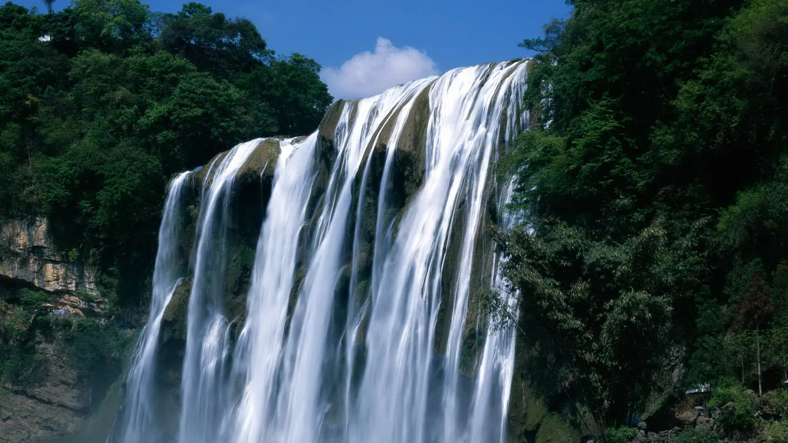 A wide waterfall cascades down a cliff face with lush green trees on either side and a blue sky above.