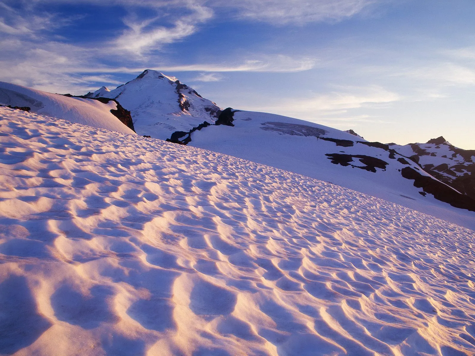 Textured snow drifts sculpted by wind cover a mountain slope, bathed in the warm glow of sunset.