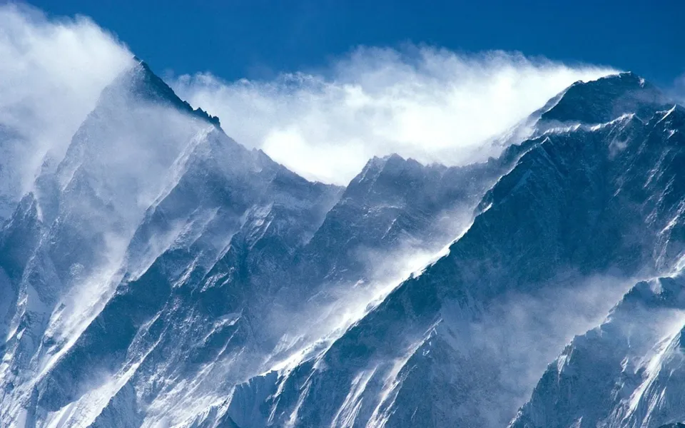 Snowy mountain peaks are buffeted by strong winds and swirling mist under a bright blue sky.