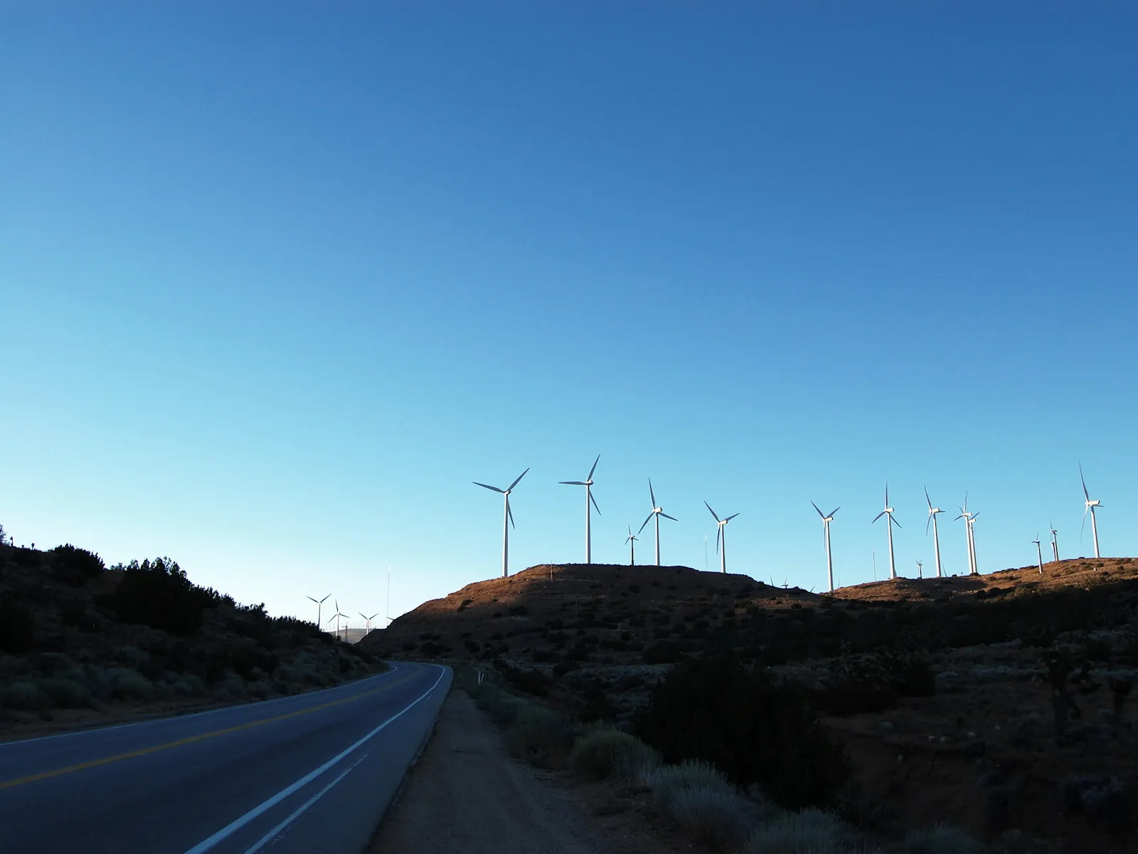 A road curves past a hillside dotted with wind turbines under a clear blue sky.
