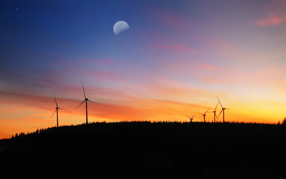 Silhouettes of wind turbines on a dark ridge under a crescent moon and colorful sunset sky.